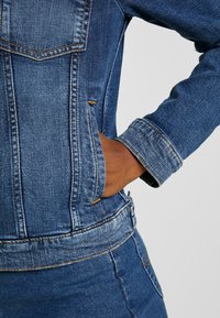 Jeansjacka med en blå tygtextur, med sömnadsdetaljer, en figursydd design och en hand stoppad i en sidoficka.