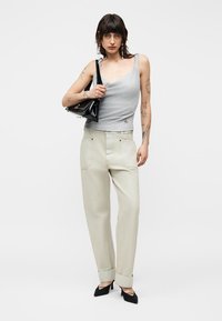 Γυναίκα με τατουάζ που φοράει γκρι αμάνικο τοπ, μπεζ παντελόνι με φαρδιά πόδια, μαύρα τακούνια και κρατάει μια μαύρη τσάντα στον ώμο της.