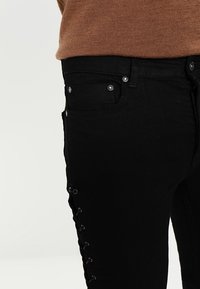 Jeans de mezclilla negra con detalle de cordones en el lateral y acentos metálicos. La tela es suave y con un ligero estiramiento para mayor comodidad.