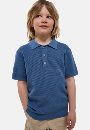 Jeune enfant aux cheveux blonds mi-longs portant un polo en maille bleu à manches courtes et un pantalon beige, debout les mains dans les poches.