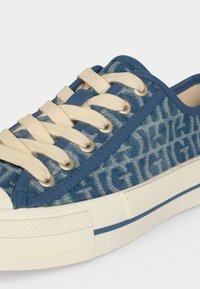 Baskets bleues avec lacets crème, semelle en caoutchouc blanche et dessus en tissu présentant un motif de texte estompé répété.