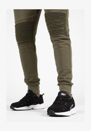 Olivengroene tapered joggers met ribbelaccenten op de dijen, gecombineerd met zwarte hardloopschoenen met een witte zool en logo-details.
