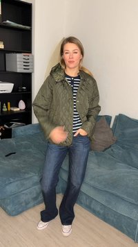 Γυναίκα με ελιά πράσινο καπιτονέ μπουφάν, ριγέ μπλε πουκάμισο, τζιν και λευκά αθλητικά παπούτσια, στέκεται σε σαλόνι με πετρόλ καναπέ και μονάδα ραφιών.