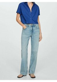 Camicia blu corta con colletto, abbinata a jeans chiari a vita alta. Il modello indossa sandali neri e ha le mani nelle tasche.