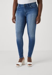 Blue denim skinny jeans with a mid-rise waist, featuring front pockets and slight fading. Paired with white open-toe block-heeled sandals.