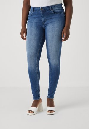 Jean skinny en denim bleu avec une taille mi-haute, comportant des poches avant et un léger délavage. Associé à des sandales à talons blocs ouvertes blanches.