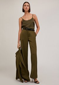 Vihreä silkki-camisole ja korkeavyötäröiset housut mustalla vyöllä. Malli pitää kädessään yhteensopivaa bleiseriä. Mustat remmikorot täydentävät asua.