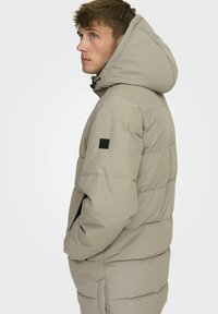 Beige Steppjacke mit Kapuze, ausgestattet mit einem gesteppten Design, seitlichen Taschen und einem schwarzen Akzent am linken Ärmel. Glatte Textur.