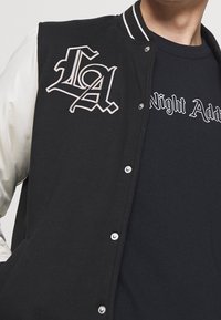 Černá varsity bunda s bílými saténovými rukávy, vyšívané logo "LA" a patentky. V kombinaci s černým tričkem s potiskem pod ní.