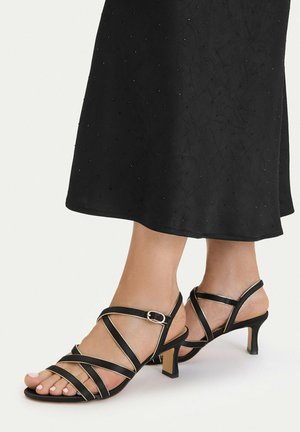 Stopy w czarnych sandałach na średnim obcasie z paskami i sprzączkami, zestawione z czarną sukienką o delikatnej kropkowanej fakturze.