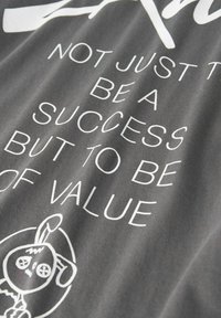 Grå stof t-shirt med hvid tekst, der står: "IKKE BARE FOR AT VÆRE EN SUCCES MEN FOR AT VÆRE AF VÆRDI," med en illustration af en tegneseriefigur.
