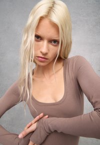 Maglietta a maniche lunghe, attillata, in tessuto a coste di un marrone chiaro. Presenta un collo a barca e un design aderente. Le maniche si estendono a coprire i polsi.