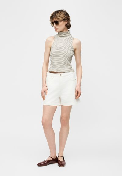 Γυναίκα με στεφάνη χωρίς μανίκια με ρίγες, λευκά σορτς, καφέ παπούτσια Mary Jane και γυαλιά ηλίου, στέκεται μπροστά σε απλό φωτεινό φόντο.