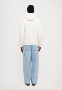 Cream hoodie with a large hood, ribbed hem, and textured fabric. Paired with loose-fitting light blue jeans and dark brown shoes.
