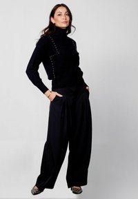 Melnā turtlenecka jaka ar studētām akcentiem, apvienota ar platām kājām melnām biksēm. Tekstūra izskatās kā adīta; apaviem ir studu detaļas.