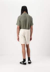 Kortærmet olivengrøn skjorte med et struktureret mønster, parret med beige stribede shorts. Model iført sorte sneakers og sokker.