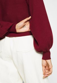 Burgundy stickad tröja med puffärmar, instoppad i midjan över vita hög midja jeans, som visar en tatuering på handen.