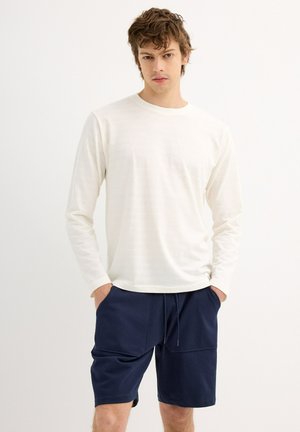 Camisa blanca de manga larga hecha de tela suave, combinada con pantalones cortos azul marino con bolsillos y cordón. Diseño simple y casual.