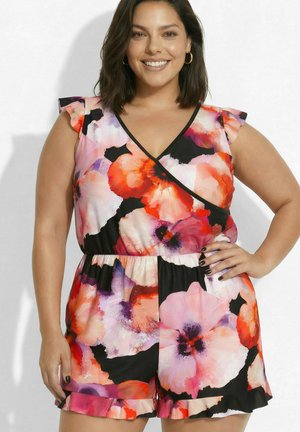 Femme souriante aux cheveux foncés mi-longs portant une combinaison fleurie sans manches avec des manches à volants et un short, main sur la hanche.