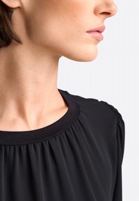 Zwarte top met een ronde halslijn en plooidetails, gemaakt van een soepele stof. Close-up weergave die de halslijn en schouder benadrukt.