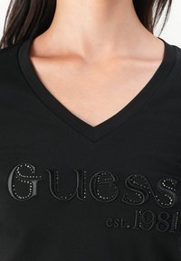 Juodas V formos marškinėliai su iškilmingu "Guess est. 1981" įspaudu ir sidabriniais kalnų krištolais, akcentuojantys glotnią audinio tekstūrą ir modernų dizainą.