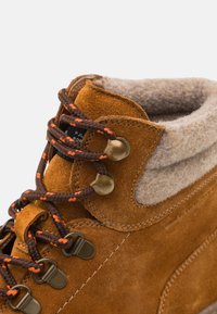 Botas de ante marrón con un cuello de lana, presentando cordones trenzados en marrón oscuro y naranja, ojales de metal y una textura suave.