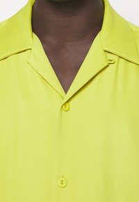 Gros plan d'une personne à la peau foncée portant une chemise jaune vif à col, avec deux boutons visibles attachés.