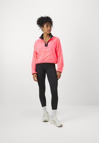 Veste courte néon rose avec col noir, fermeture éclair mi-hauteur et poche avant, associée à des leggings noirs avec des accents texturés et des baskets blanches.