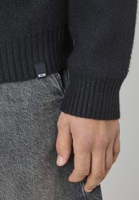 Pull noir en tricot avec des poignets côtelés et une petite étiquette BZB à l'ourlet. Associé à un pantalon gris texturé, mains visibles.