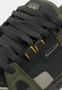 Gray sneaker with green accents, featuring a textured upper, thick gray laces, and a yellow accent on the lacing area.
