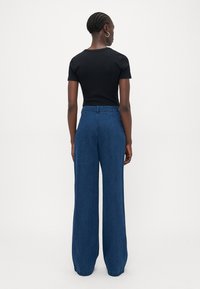 Ženska z pletenicami, ki nosi srebrne obročaste uhane, črno kratkotrajno majico in svetlo modre široke hlače z visokim pasom, stoji ob beli steni.