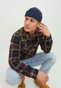 Camisa de franela a cuadros en azul marino, rojo y beige, pantalones vaqueros azul claro, botas marrones y un gorro de punto azul marino; pose sentado con la mano en la cabeza.