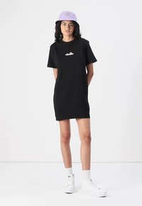 Juodasis medvilnės T-shirt su trumpomis rankovėmis, su balta logotipu ant krūtinės. Suderintas su šviesiai purpurine kibiru ir baltais sportbačiais.