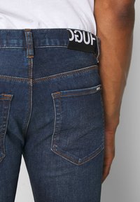 Modré denimové džíny s přiléhavým střihem, viditelným šitím a dvěma zadními kapsami. Na pásu se nachází černá nálepka s bílým textem.