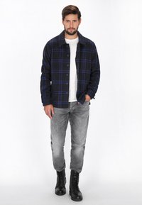 Blauwe en zwarte geruite shirtjack, met knoopsluiting, iets lose fit, gecombineerd met grijze jeans en zwarte veterschoenen.