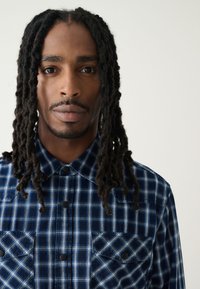 Chemise à carreaux bleu marine et bleu clair avec deux poches poitrine et boutons pression noirs, fabriquée dans un tissu texturé.