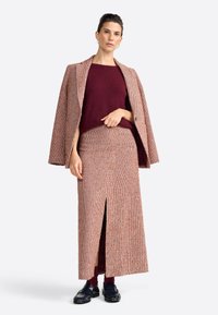 Textured, check-patterned coat in shades of red and beige, paired with a burgundy knit top and coordinating long skirt with a front slit.