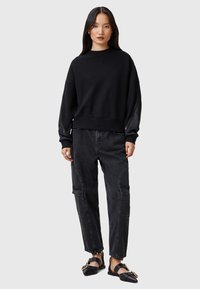 Felpa nera oversize con spalle scese, abbinata a pantaloni cargo grigi scuri con tasche e dettagli sulle cuciture. Sandali neri con accenti dorati.