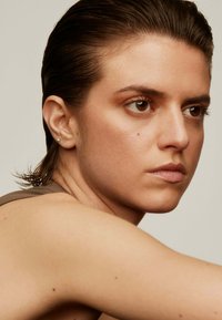 Primer plano de una persona con pelo corto y mojado, vistiendo una camiseta sin mangas marrón. Las características incluyen una textura de piel suave, ojos marrones y pendientes pequeños.