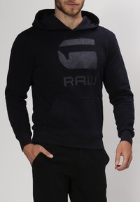 Felpa con cappuccio blu navy con un grande logo testurizzato e la scritta "RAW" in un colore a contrasto. Presenta una tasca frontale e polsini a costine. Tessuto morbido.