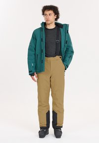 Teal waterdichte jas met ritsen, zwarte accenten; beige broek met zwarte zijpanelen en verstelbare banden; gedragen met zwarte schoeisel.