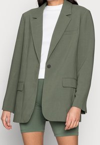 Olivegrüner, übergroßer Blazer mit einem Ein-Knopf-Verschluss, Reverskragen und zwei vorderen Taschen, kombiniert mit einem weißen Hemd und grünen Shorts.