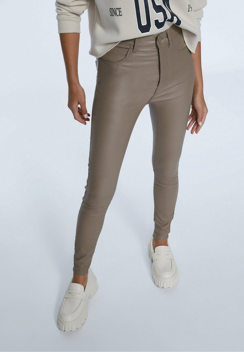 Femme portant un pantalon skinny taille haute taupe et des mocassins blancs épais, associés à un sweatshirt beige avec du texte noir sur les manches.