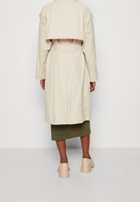 Persona vistiendo un abrigo trench beige con cinturón, una falda verde oliva y zapatos de tacón grueso beige, de pie contra un fondo claro y liso.