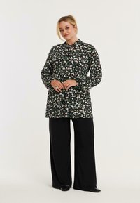 Zwarte bloemenblouse met een geknoopte kraag, lange mouwen en een losse pasvorm, gecombineerd met zwarte wijde broek en donkere schoenen.