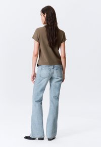 Olivno zelena bombažna majica s kratkimi rokavi in rahlo krajši kroj, kombinirana s svetlo modrimi širokimi kavbojkami in črimi čevlji.