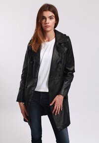 Cappotto trench in pelle nera con design doppiopetto, spalline e tasche laterali, indossato sopra una camicia bianca e jeans.