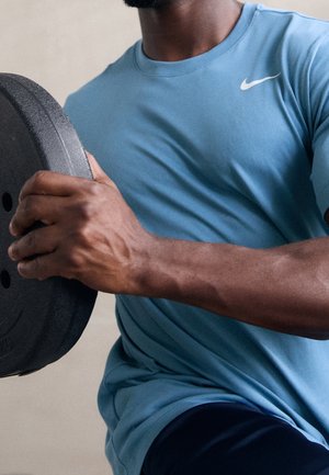 Mann im blauen Hemd hält eine schwarze Gewichtsscheibe und zeigt während des Krafttrainings seinen muskulösen Arm und Brust.