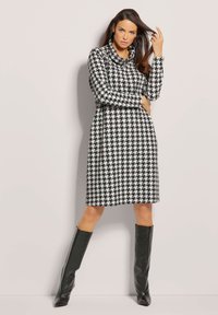 Langarmkleid im Hahnent pattern, aus schwarz-weißer Stoff gefertigt. Es hat einen Cowl-Ausschnitt und wird mit knielangen schwarzen Stiefeln kombiniert.