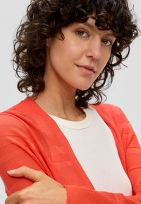 Cardigan corail avec motif de rayures texturées, superposé sur un t-shirt blanc. Tissu doux, design à devant ouvert et coupe décontractée.